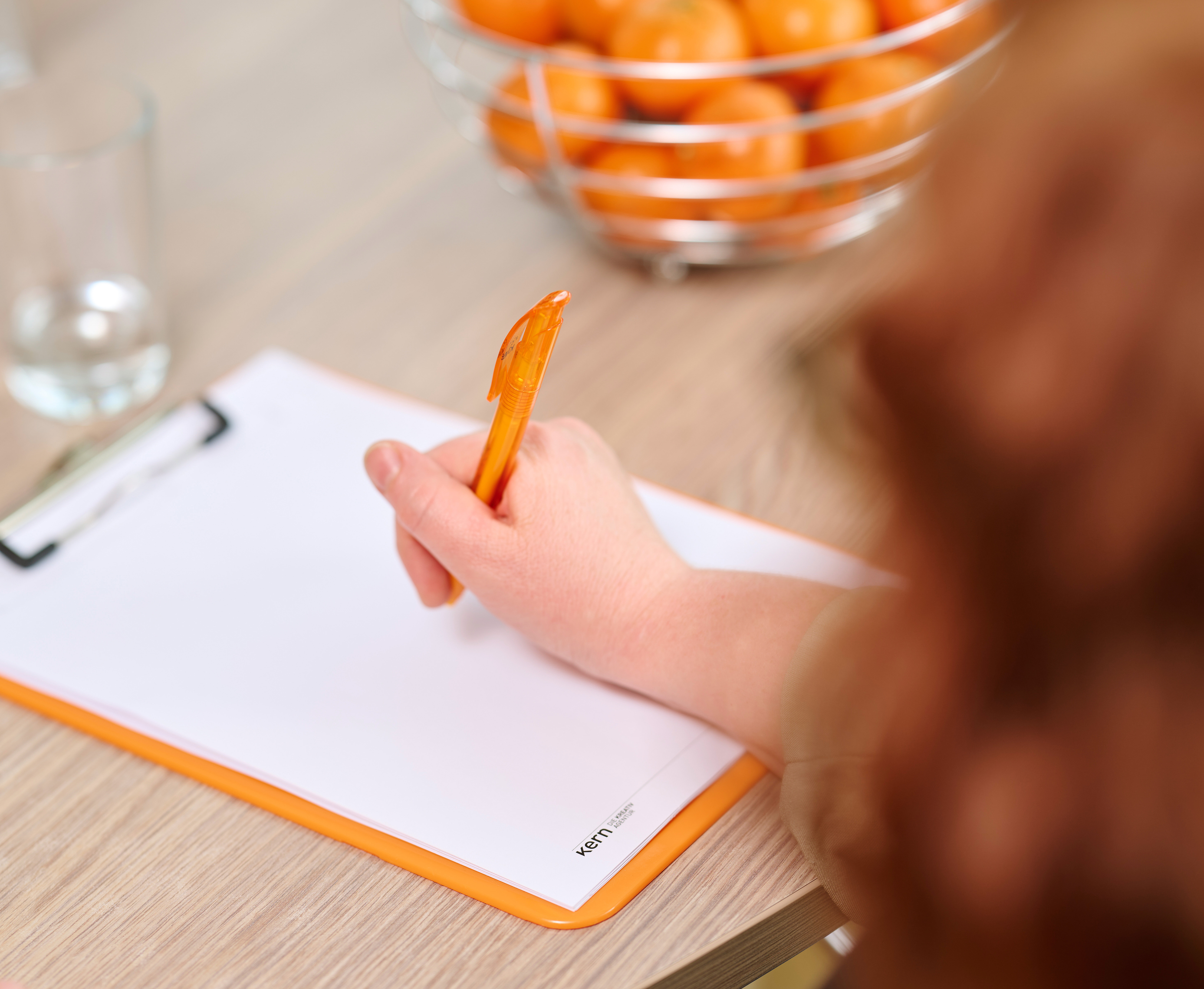 Schulterblick von Hand mit orangenem Kugelschreiber macht sich auf einem Klemmbrett notizen